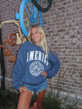 Load image into Gallery viewer, Person wearing a blue sweatshirt with 'America' printed on it, lying on the ground.