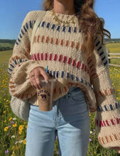 Load image into Gallery viewer, A woman wearing a chunky knit sweater with a cream base and colorful horizontal stripes in blue, red, and brown, standing in a flower field and holding a coffee cup.