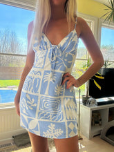 Load image into Gallery viewer, Woman wearing a blue and white patterned dress standing by a window with a view of a pool.