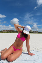 Load image into Gallery viewer, Woman in pink bikini and white cap on a beach with blue sky and clouds.