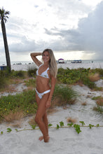Load image into Gallery viewer, Woman in a white bikini lying on a sandy beach with grass and palm trees.