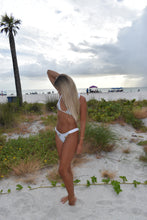 Load image into Gallery viewer, Woman in a white bikini standing on a beach with palm trees and sand dunes in the background.