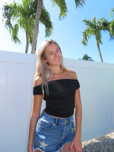 Load image into Gallery viewer, A female model wearing an off-the-shoulder black crop top and denim shorts, standing by a fence with palm trees in the background.