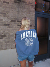 Load image into Gallery viewer, Person wearing a blue sweatshirt with 'AMERICA' printed on it, standing on a brick pavement.