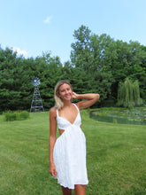 Load image into Gallery viewer, Woman in a white dress standing in a grassy field with trees and a windmill in the background.