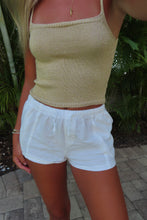 Load image into Gallery viewer, A woman wearing a beige ribbed knit camisole top and white shorts, standing outdoors on a paved walkway with green foliage in the background.