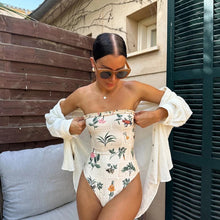 Load image into Gallery viewer, A strapless one-piece swimsuit with a floral pattern, worn by a person standing in front of a green shuttered window.
