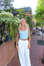 Load image into Gallery viewer, Woman in a white dress standing on a sidewalk with greenery and people in the background