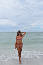 Load image into Gallery viewer, Woman in a pink bikini standing on a beach with ocean waves.