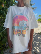 Load image into Gallery viewer, A person wearing a white t-shirt with a vibrant pink sunset and beach elements design, standing on a beach.