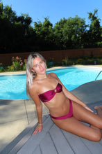 Load image into Gallery viewer, A woman wearing a magenta satin bikini sits on the edge of a swimming pool outdoors, with trees and a wooden fence in the background.