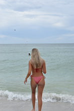 Load image into Gallery viewer, Woman in a pink bikini walking along a beach with clear blue water and white waves.