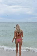 Load image into Gallery viewer, Woman in a pink bikini walking along a beach with clear blue water and white waves.