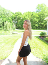 Load image into Gallery viewer, Woman in a white top and black skirt standing outdoors with greenery in the background