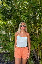 Load image into Gallery viewer, A woman with blonde hair and sunglasses stands in front of lush green palm leaves, wearing a white knit strapless tube top and peach-pink shorts.
