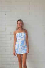 Load image into Gallery viewer, Woman wearing a strapless dress with blue abstract pattern against a white brick wall.