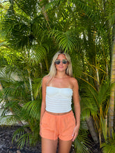 Load image into Gallery viewer, A woman stands outdoors in front of lush green palm foliage, wearing a white strapless crochet tube top and peach-orange drawstring shorts.
