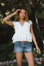 Load image into Gallery viewer, Woman wearing a white blouse and denim shorts standing outdoors with trees in the background
