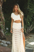 Load image into Gallery viewer, A woman stands outdoors in front of trees wearing a cream two-piece boho-style outfit consisting of a ruffled short-sleeve crop top and a matching high-slit maxi skirt with an openwork crochet pattern.
