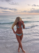 Load image into Gallery viewer, Woman in a bikini standing on a beach with ocean waves and sunset sky.
