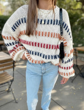 Load image into Gallery viewer, A woman wearing a chunky knit sweater with horizontal stripes in navy blue, terracotta orange, and red on an off-white base, paired with light-wash blue jeans in an outdoor patio setting.

