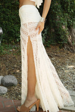 Load image into Gallery viewer, A woman outdoors wearing a long white lace maxi skirt with a high thigh slit and a matching white crop top. The background shows green foliage, trees, and a stone path.
