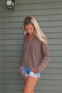 Woman wearing a brown knitted sweater and denim shorts standing against a gray wall.