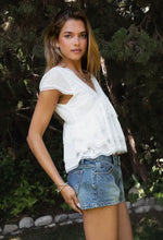 Load image into Gallery viewer, Woman wearing a white top and denim shorts standing outdoors with trees in the background
