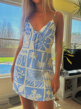 Load image into Gallery viewer, Woman wearing a blue and white patterned dress standing indoors near a window with a view of a pool.
