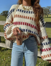 Load image into Gallery viewer, A person outdoors wearing a chunky knit sweater with horizontal multicolor stripes in cream, rust orange, and navy blue, paired with light blue jeans and holding an iced coffee.
