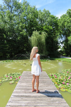 Load image into Gallery viewer, Woman in a white dress standing on a wooden dock by a pond with lily pads and trees in the background.
