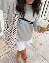 Load image into Gallery viewer, A woman wearing a light gray knit sweater with a blue and black fish graphic on the front, styled with a white ruffled mini skirt, tan ankle boots, and a black-and-white polka dot tote bag. She is holding a green drink on a sidewalk.
