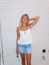 Load image into Gallery viewer, Woman lying on a bed wearing a white top and denim shorts, with sunglasses beside her.
