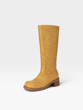 Load image into Gallery viewer, A single knee-high boot in a warm mustard yellow faux-leather material, featuring a low brown block heel and a rounded toe, shown against a plain light gray background.
