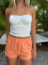 Load image into Gallery viewer, A woman wearing a white crocheted strapless tube top and peach-colored shorts, standing outdoors in front of a hammock and palm trees.

