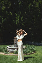 Load image into Gallery viewer, A woman posing outdoors in a garden setting with a stone fountain and lush green trees. She is wearing a matching white crochet two-piece set consisting of a ruffled short-sleeve crop top and a long skirt.
