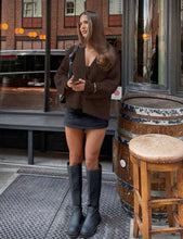 Load image into Gallery viewer, Woman in a brown coat and black boots standing outside a building with wooden barrels and stools.
