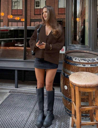Woman in a brown coat and black boots standing outside a building with wooden barrels and stools.