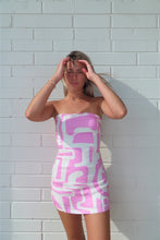 Load image into Gallery viewer, Woman wearing a pink and white patterned dress against a light gray brick wall.
