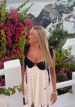 Load image into Gallery viewer, Woman in a black and white outfit standing in front of pink flowers and a scenic background.
