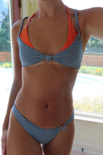 Load image into Gallery viewer, A woman wearing a two-piece bikini with a blue and white gingham pattern and bright orange inner cups, standing in front of a window.
