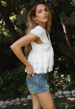Load image into Gallery viewer, Woman in a white top and denim shorts standing outdoors with trees in the background
