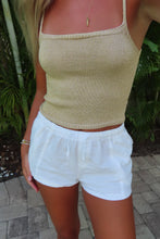 Load image into Gallery viewer, A woman wearing a beige ribbed knit camisole top and white drawstring shorts, standing outdoors on a paved walkway with green plants in the background.
