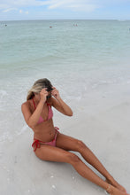 Load image into Gallery viewer, Woman in a red bikini lying on a sandy beach with clear blue water.
