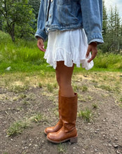 Load image into Gallery viewer, A person outdoors wearing a white ruffled mini skirt, a light blue denim jacket, and brown knee-high leather boots on a dirt path with grass and trees in the background.
