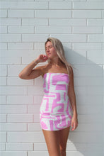 Load image into Gallery viewer, Woman wearing a pink and white patterned dress against a white brick wall.

