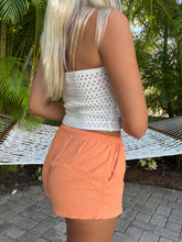 Load image into Gallery viewer, A woman shown from behind wearing a white crochet knit crop top and peach-colored shorts, standing outdoors on a paved surface with palm trees and a white hammock in the background.
