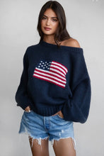 Load image into Gallery viewer, A woman wearing a navy blue off-the-shoulder knit sweater with a large American flag motif on the front, paired with distressed denim shorts, against a plain light gray background.
