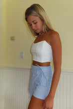 Load image into Gallery viewer, Woman wearing a white top and blue shorts against a plain wall.

