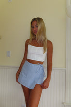 Load image into Gallery viewer, Woman wearing a white top and blue shorts standing against a yellow wall.
